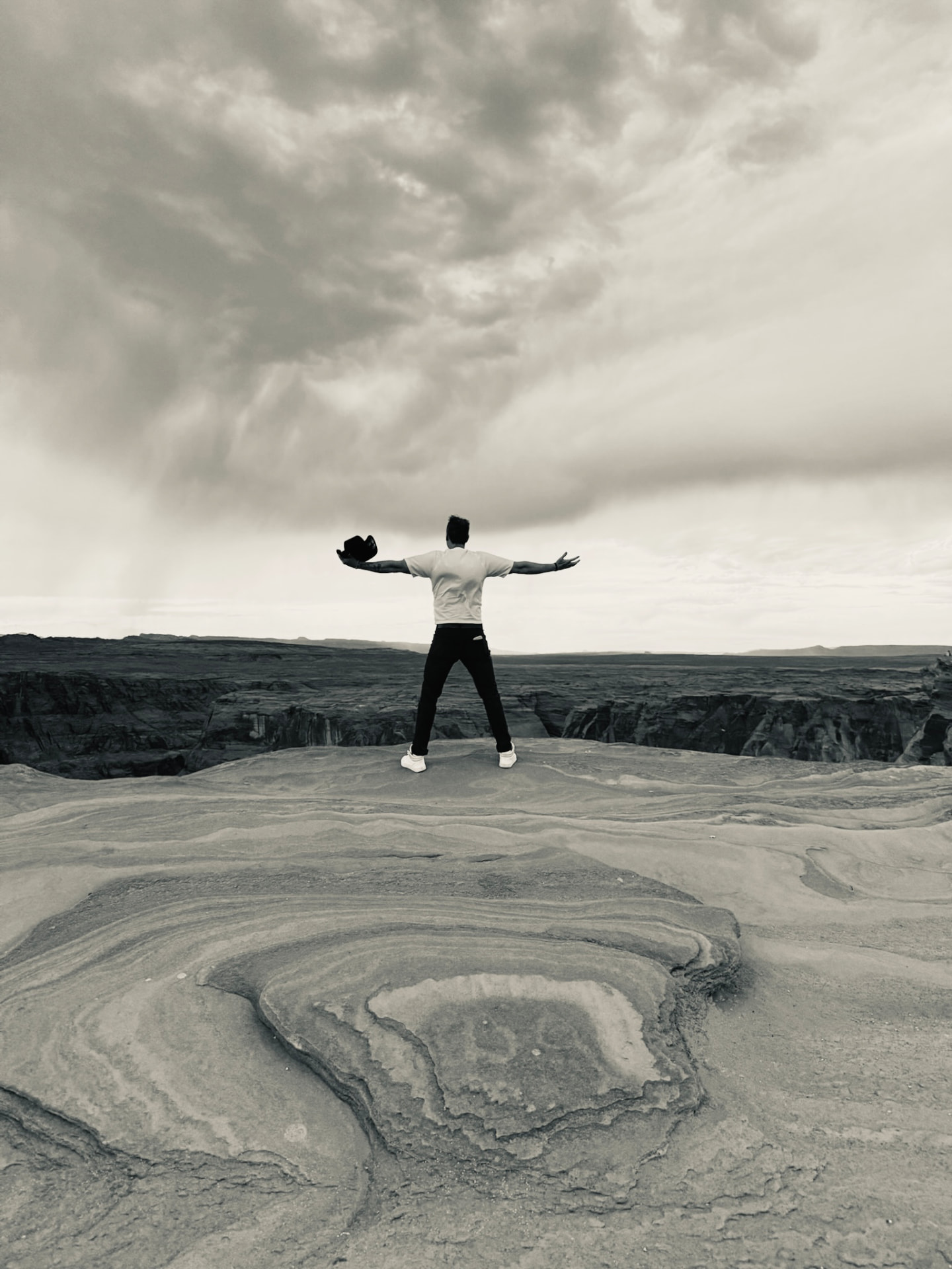 A person standing with arms open on a layered rock overlook beneath a wide, clouded sky